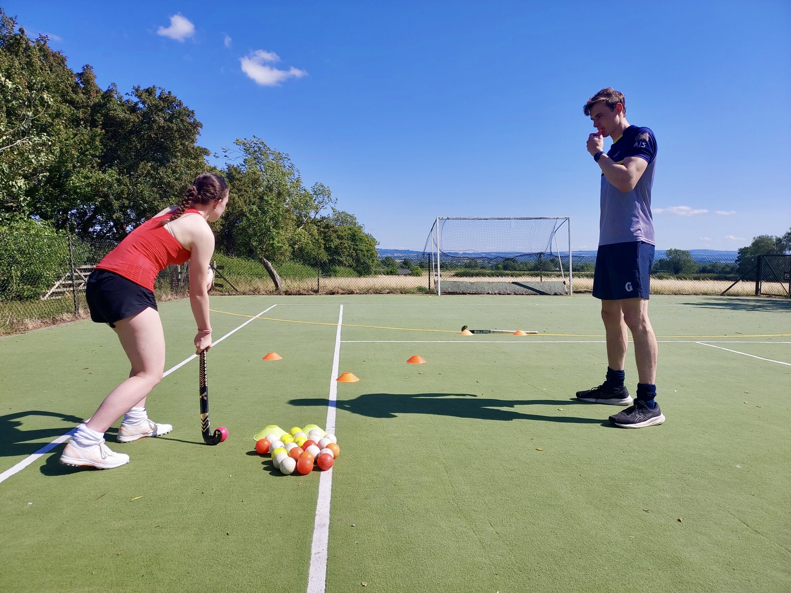 Hockey dribbling drill – skill development coaching Young hockey player preparing to start a dribbling drill around cones, with coach observing closely on the pitch with a whistle